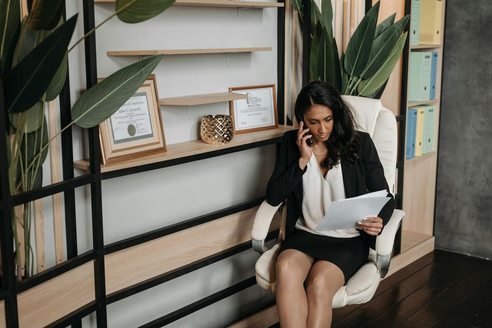 A professional woman in business attire reviews documents while on a phone call in a modern office.